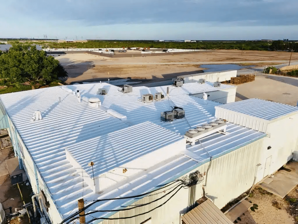 Aerial drone shot of a large commercial metal building with a freshly restored bright-white silicone-coated standing-seam roof. Multiple HVAC units, curbs, and penetrations are visible on the pristine reflective surface under a partly cloudy Texas sky.